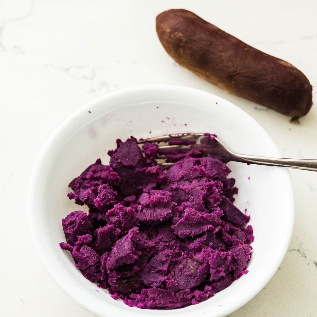 Bowl of mashed purple sweet potato with a fork, next to a whole purple sweet potato