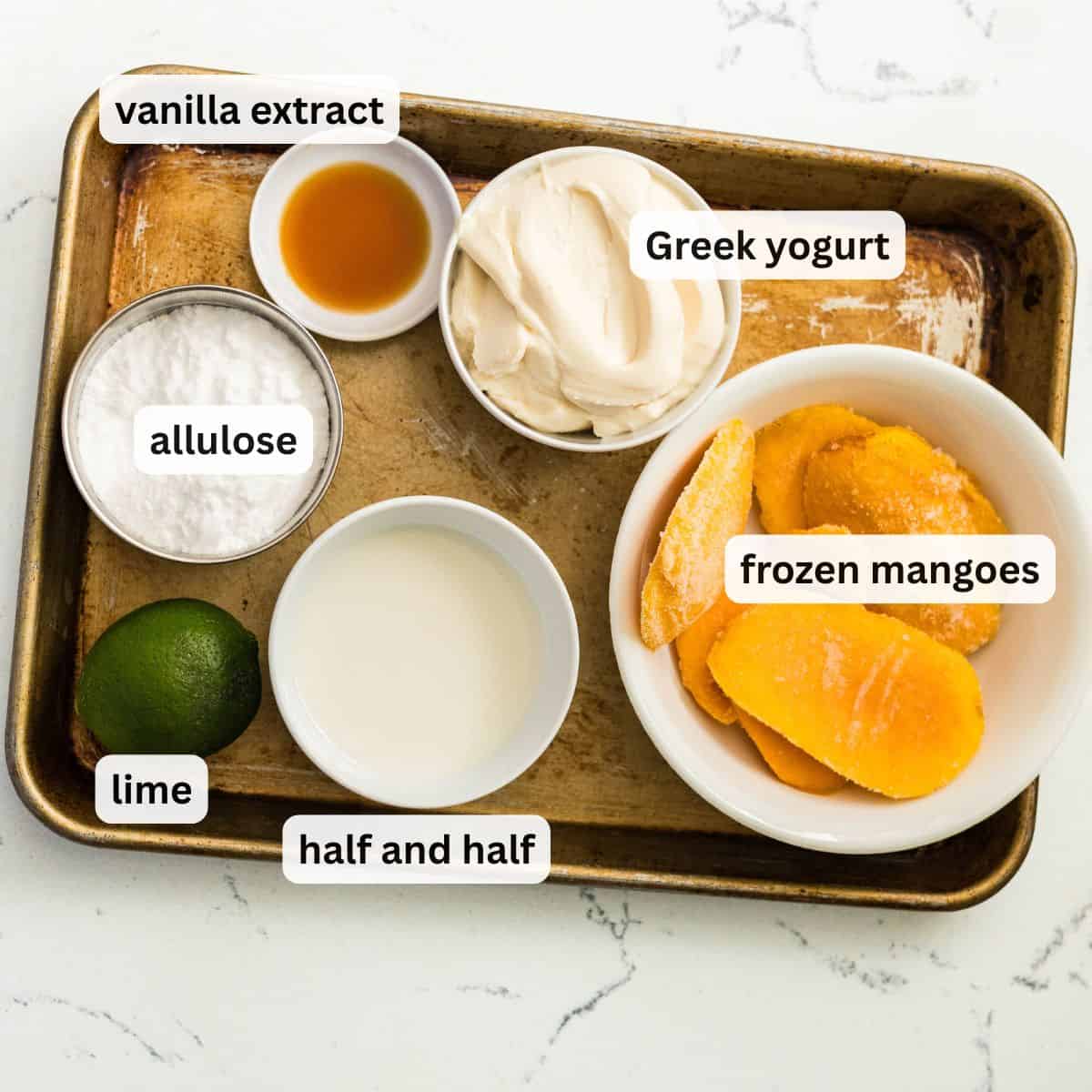 Ingredients for Ninja Creami mango frozen yogurt on a baking tray: frozen mangoes, Greek yogurt, half and half, allulose, lime, and vanilla extract